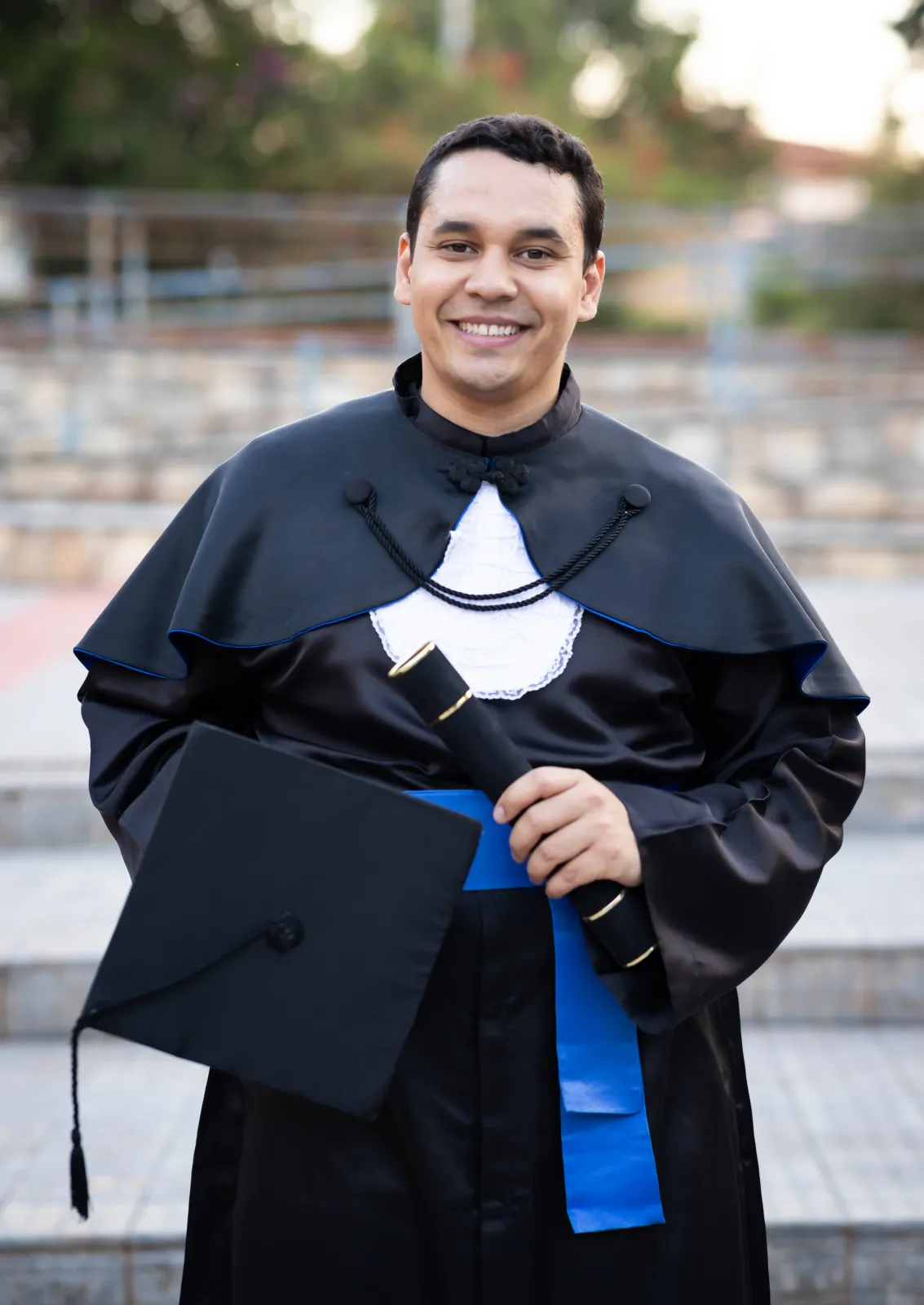 Eduardo Valente de beca, com o capelo na mão, sorrindo na escadaria do campus